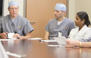Two doctors in blue surgical caps and one doctor in a white coat having a discussion at a table